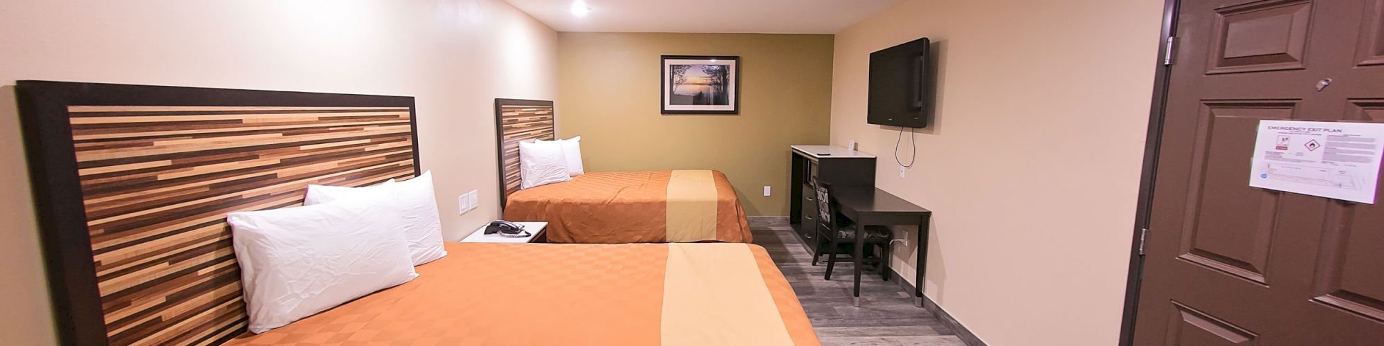 The image shows a hotel room with two double beds, a wall-mounted TV, a desk, and a brown door. The lighting is warm, enhancing the cozy ambiance.