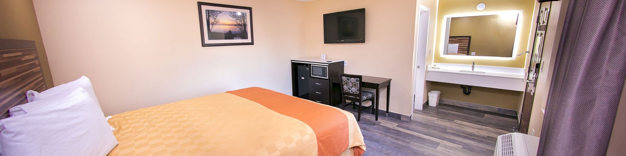 A cozy hotel room features a bed with orange bedding, a TV, a desk, and a well-lit vanity area with a mirror on the right side.