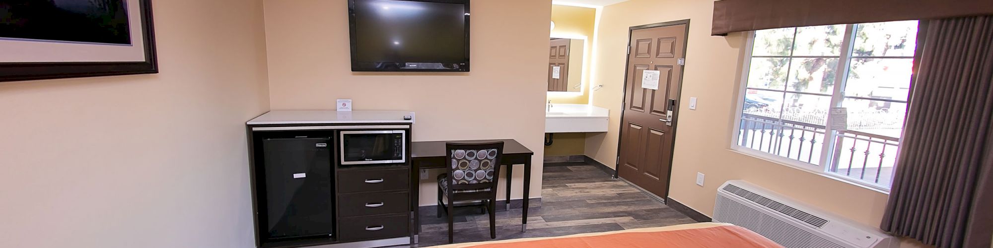 A hotel room with a bed, a wall-mounted TV, a desk, a microwave, a picture frame, a bathroom in the background, and a window with curtains.