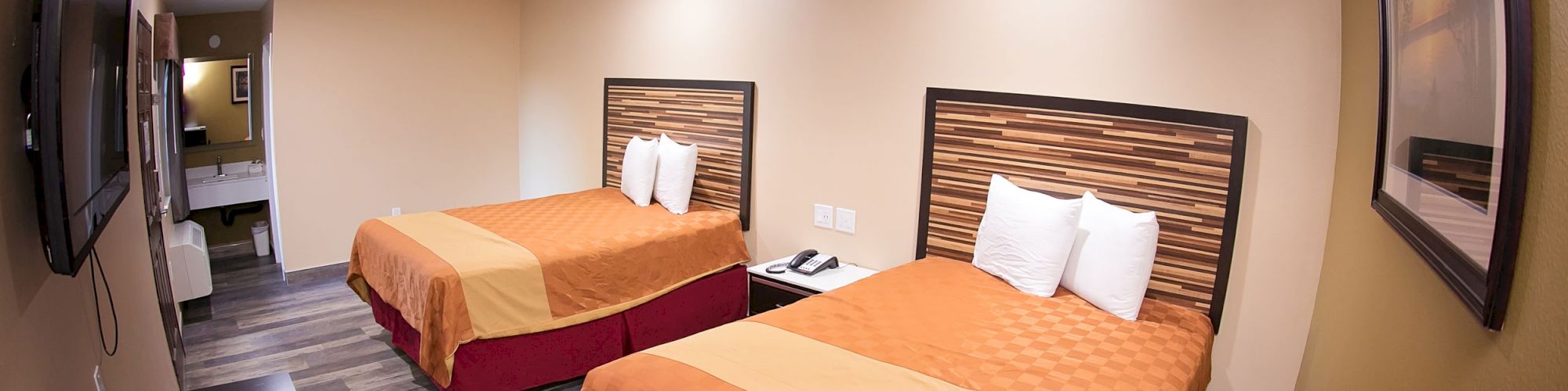 This image shows a hotel room with two beds, orange and red bedding, a TV, a table, wall art, and a bathroom visible in the background.