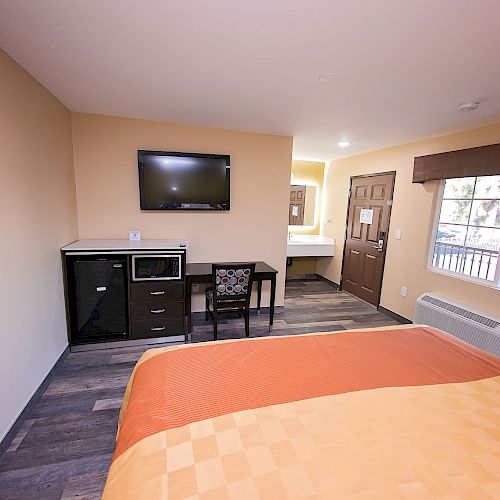 A hotel room with a bed, TV, desk, chair, microwave, and a window with curtains. There's a painting on the wall and a door in view.