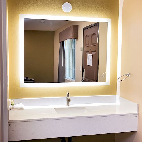 A modern bathroom with a backlit mirror, sink, and faucet. There is a towel on the counter and part of a room can be seen in the mirror's reflection.