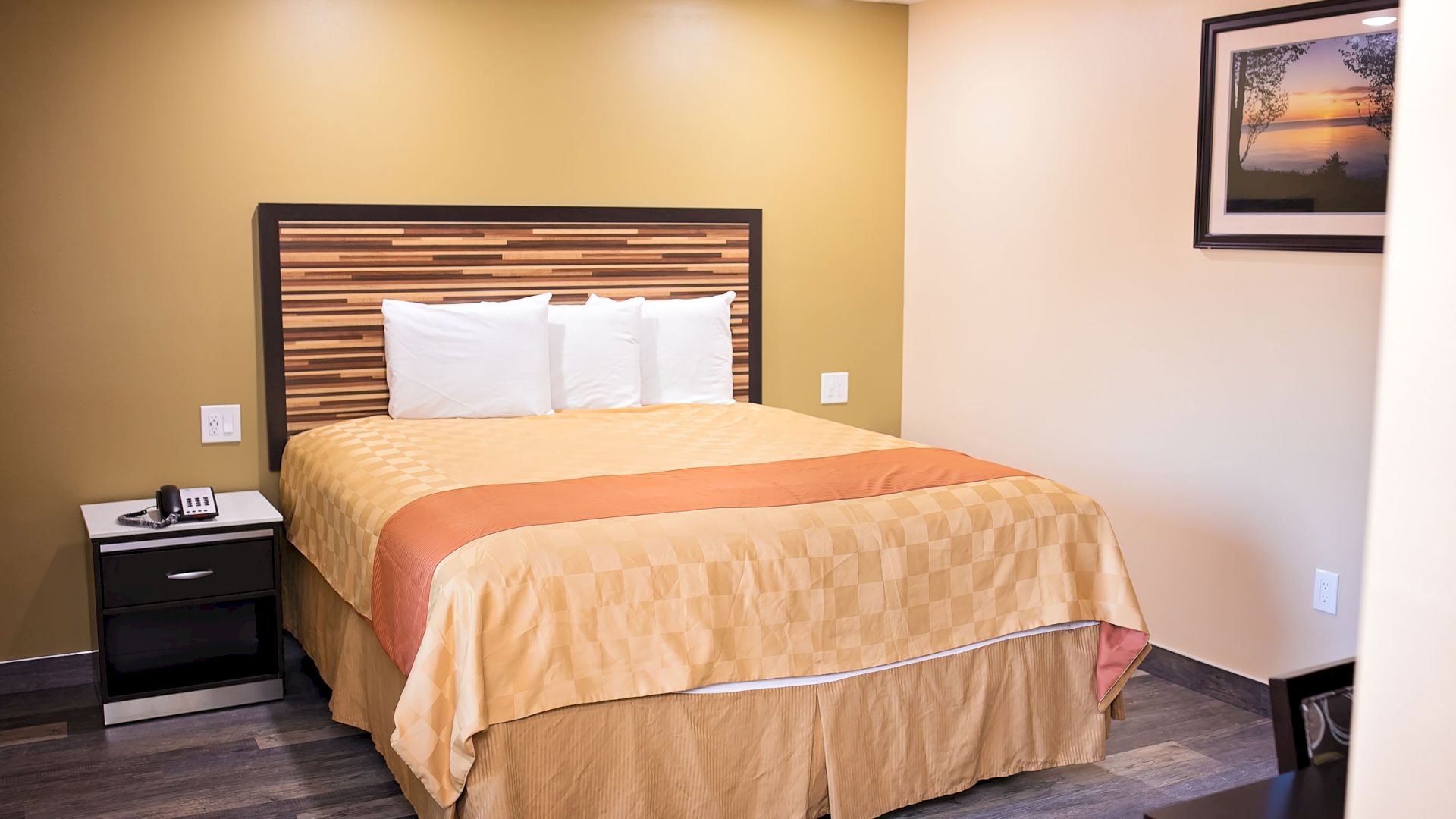 A cozy hotel room features a double bed with a patterned headboard, bedside table with a phone, and a framed picture on the wall.
