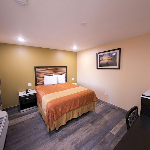 A neatly arranged hotel room features a bed, nightstand, desk, chair, and wall art, with a window providing natural light.