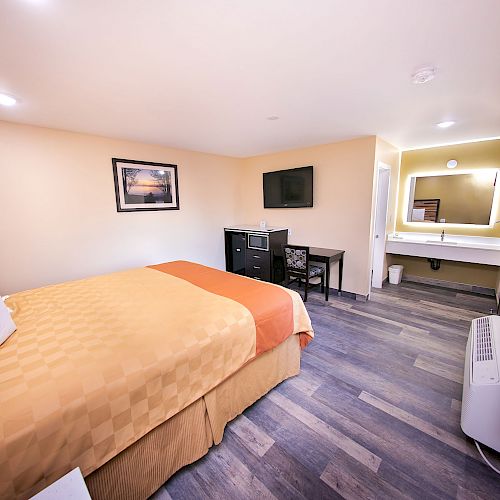 A modern hotel room featuring a bed with yellow covers, wall-mounted TV, desk, chair, mini-fridge, and a bathroom area with a lit mirror.