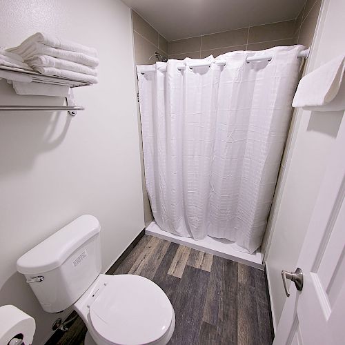 A small bathroom with a toilet, a shower with a curtain, a shelf with folded towels, and a door with a towel hanging on the back.