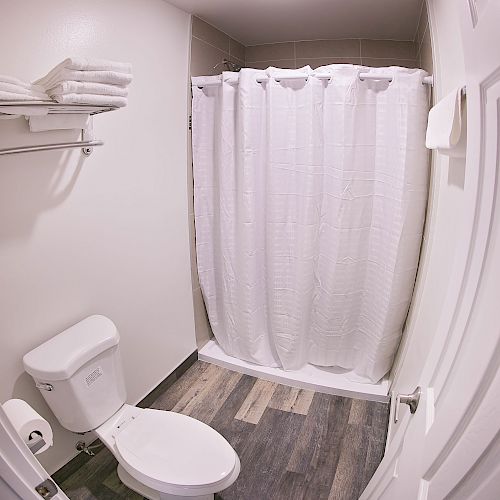 A small, clean bathroom with a toilet, shower with a white curtain, shelf with folded towels, and a towel hanging on the wall.