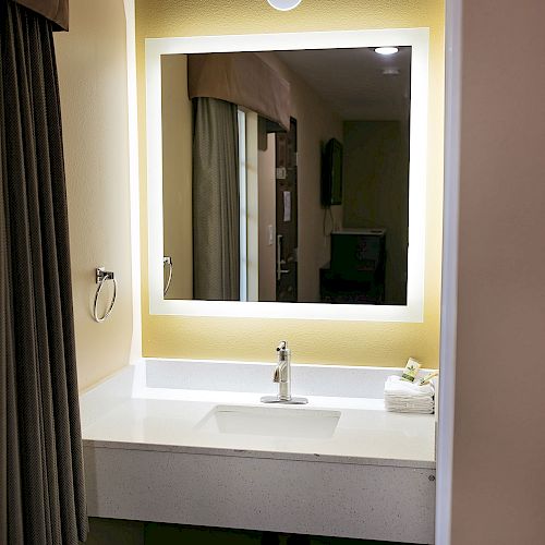 The image shows a bathroom vanity with a sink, a large illuminated mirror, toiletries, and a trash bin below, all against a warm beige wall.