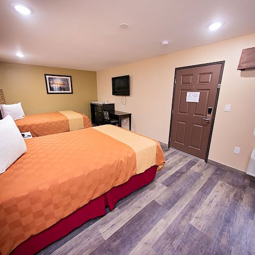 A hotel room with two beds, a TV, a desk, a window, and a door. The room is well-lit and has wooden flooring.