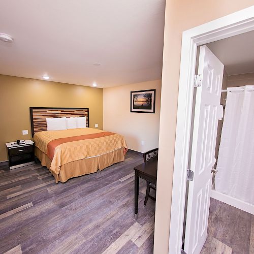 The image shows a hotel room with a bed, nightstands, a small table with a chair, and a visible bathroom with a shower curtain.