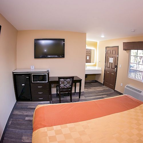 A cozy hotel room with a bed, TV, microwave, fridge, desk, and a framed photo. A door and window are visible, leading to a balcony.