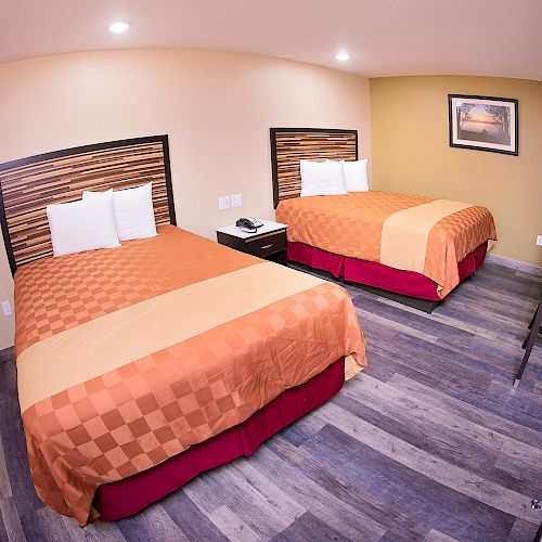 A hotel room with two double beds, a wall-mounted TV, a desk, chair, and simple decor, all in neutral tones with wooden flooring, is shown.