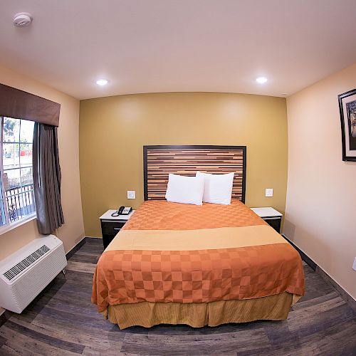 This image shows a neatly arranged hotel room with a bed, nightstands, window with curtains, air conditioning unit, and a framed picture on the wall.