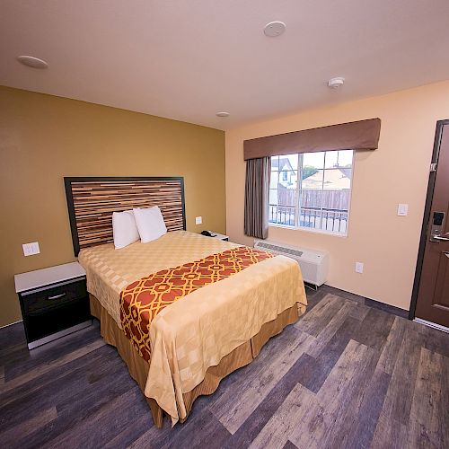 A cozy hotel room with a double bed, neatly made with a patterned bed cover, a window, and a door, wooden flooring and nightstands.