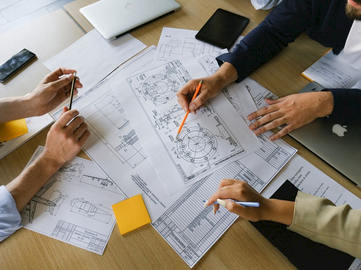 Four people collaborate over architectural plans and blueprints on a table, surrounded by notebooks, pencils, and electronic devices.