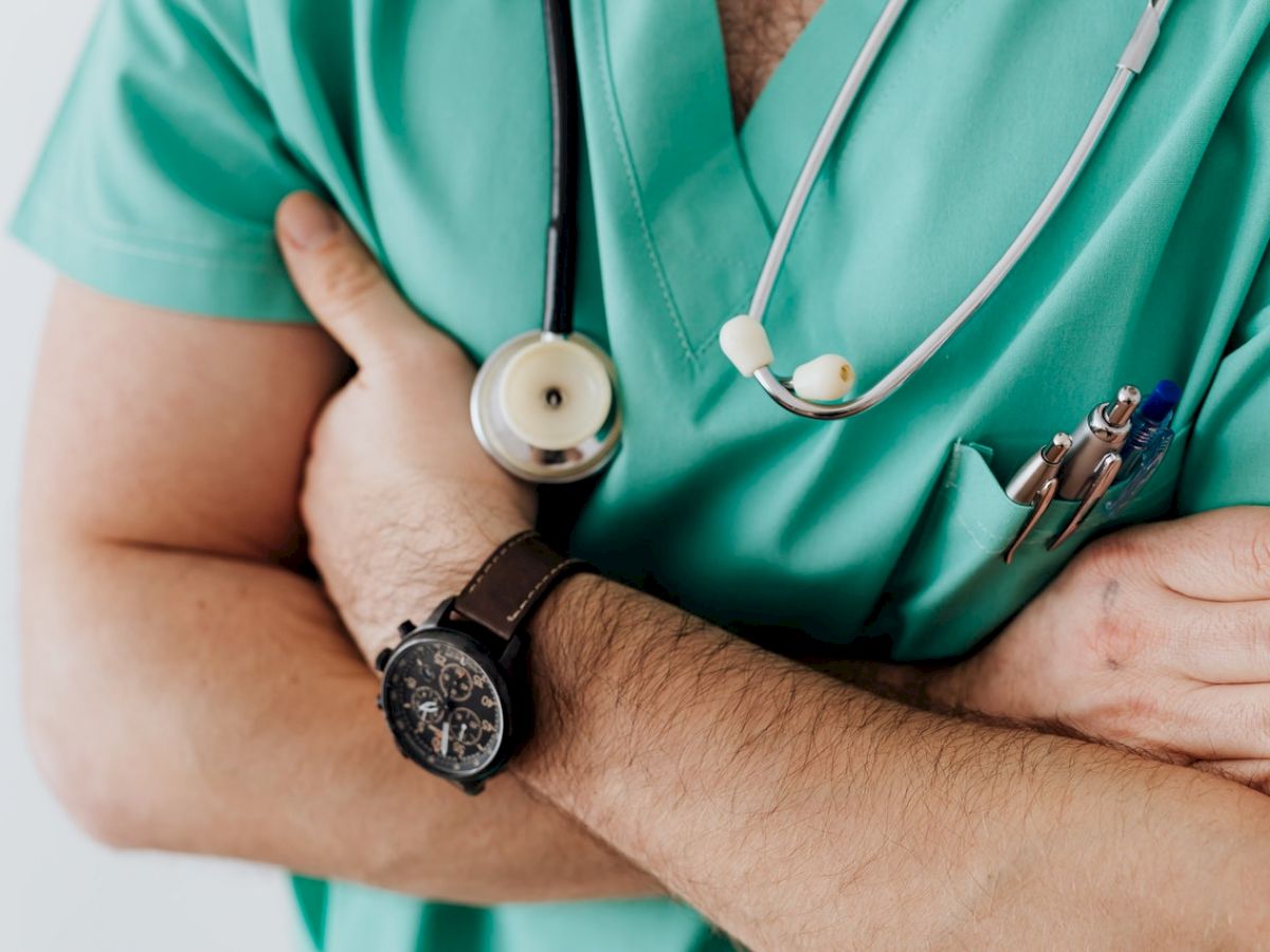 A person in green medical scrubs stands with arms crossed, wearing a stethoscope, watch, and pens in the pocket.