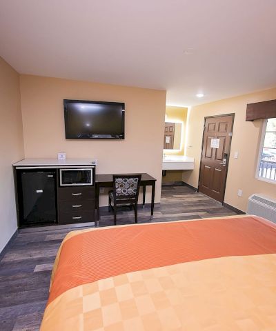 A tidy hotel room with a bed, wall TV, desk, microwave, and window with drapes. Warm lighting and neutral tones create a cozy atmosphere.