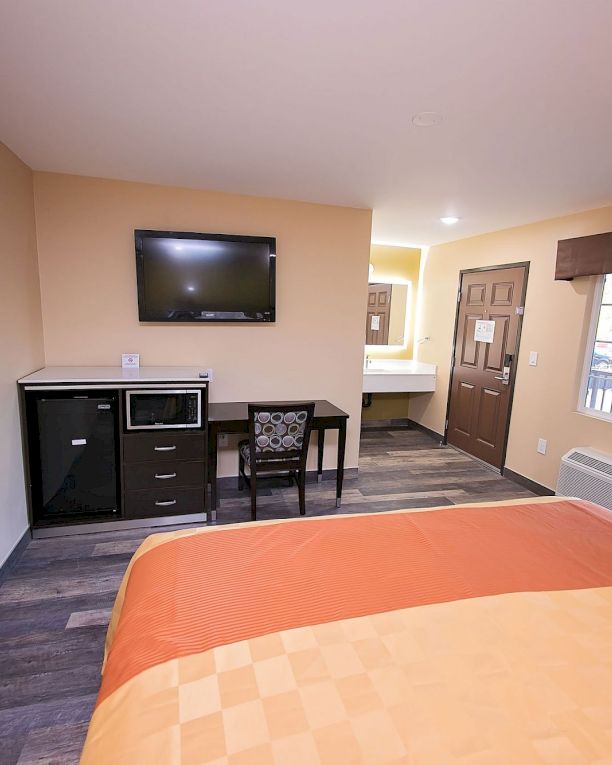 A tidy hotel room with a bed, wall TV, desk, microwave, and window with drapes. Warm lighting and neutral tones create a cozy atmosphere.