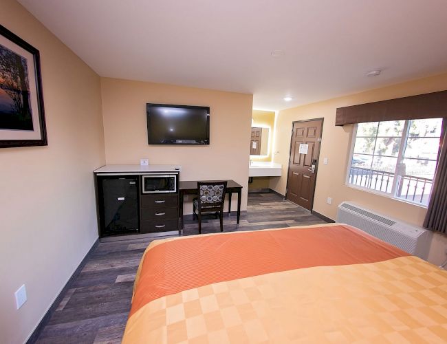 A tidy hotel room with a bed, wall TV, desk, microwave, and window with drapes. Warm lighting and neutral tones create a cozy atmosphere.