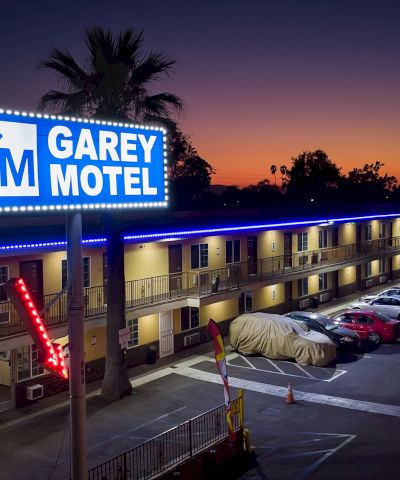 This image shows a night view of Garey Motel, with a lit sign, parked cars, and palm trees in the background against a twilight sky.