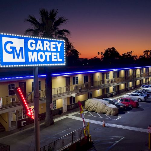 This image shows a night view of Garey Motel, with a lit sign, parked cars, and palm trees in the background against a twilight sky.