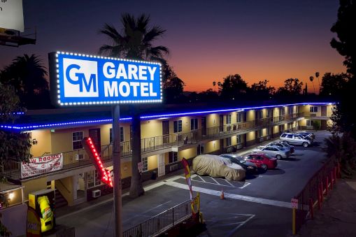 This image shows a night view of Garey Motel, with a lit sign, parked cars, and palm trees in the background against a twilight sky.