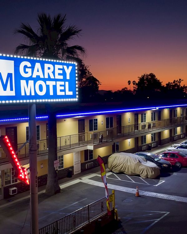 This image shows a night view of Garey Motel, with a lit sign, parked cars, and palm trees in the background against a twilight sky.