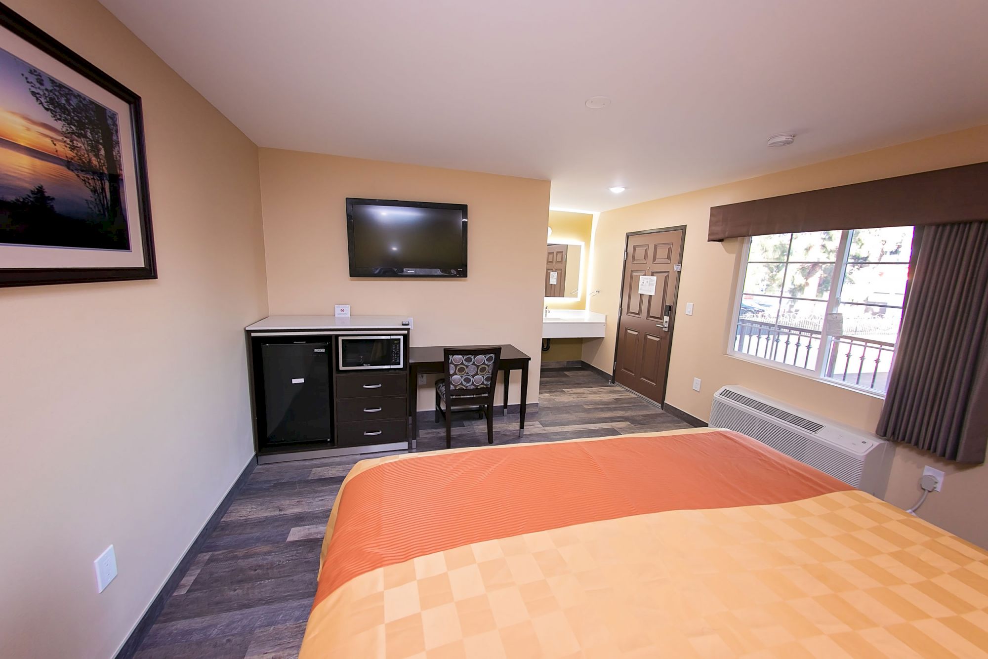 A hotel room with a bed, TV, mini-fridge, microwave, desk, chair, window, and a picture on the wall. The door leads to another area.