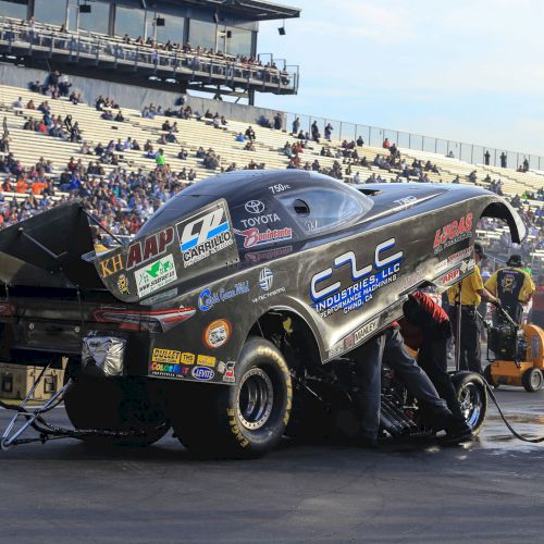 A crew works on a drag racing car with its rear lifted during a pit stop at a racing event, with spectators visible in the background.