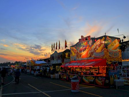 A fairground or carnival at dusk, with food stalls and colorful signs, including one for hot dogs. The sky features a beautiful sunset.