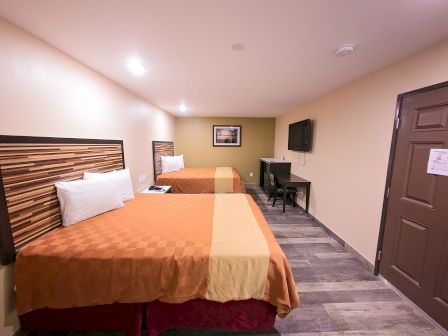 The image shows a hotel room with two double beds, a wall-mounted TV, a desk, a chair, and a door, all set in a well-lit space with simple decor.