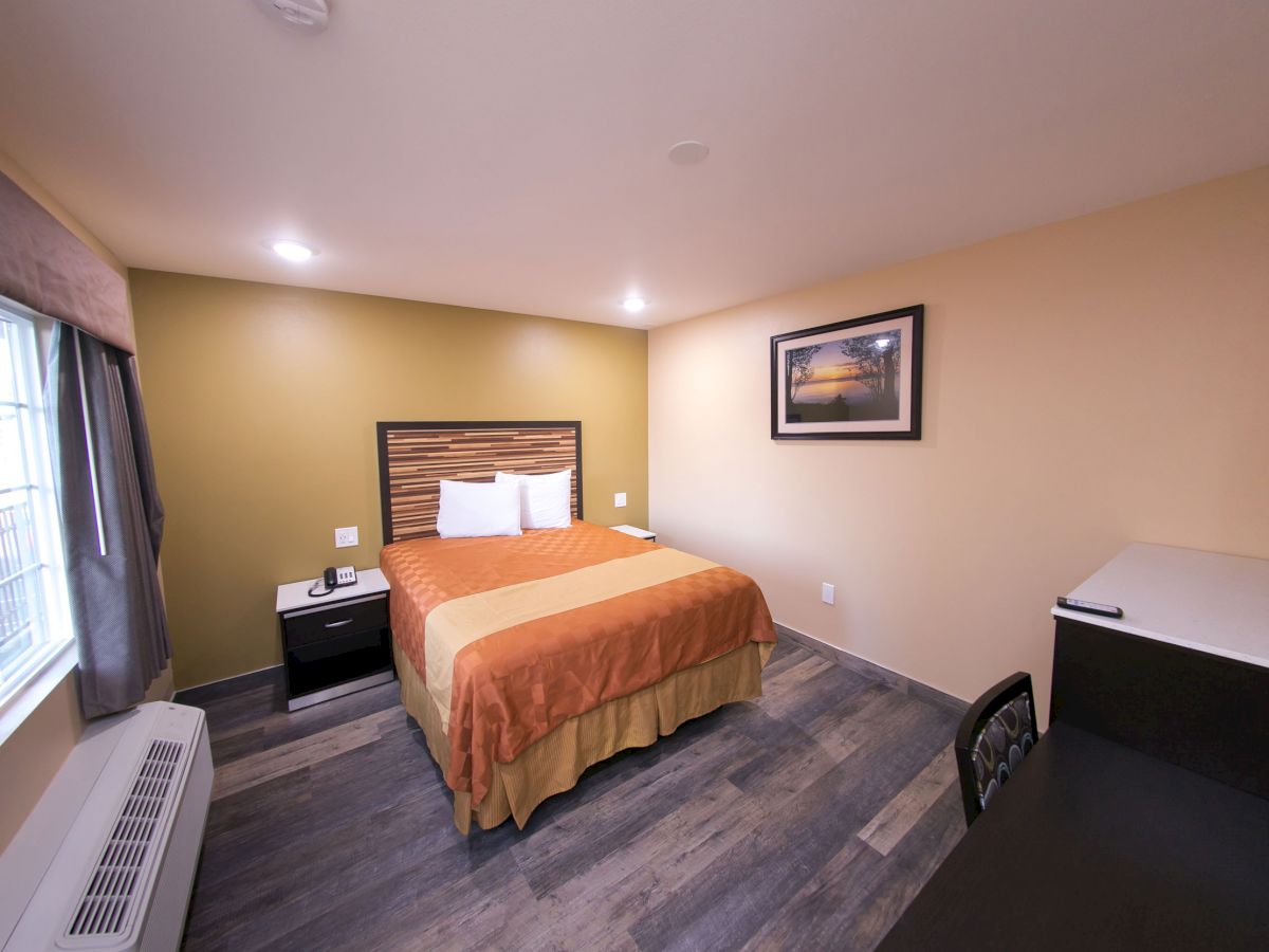 A cozy hotel room with a double bed, side table, window, desk, and framed picture on the wall, featuring minimalist decor and warm lighting.