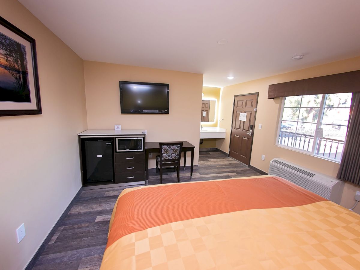 A hotel room with a bed, TV, desk, chair, microwave, and fridge. There's a window, door, and a picture on the wall.